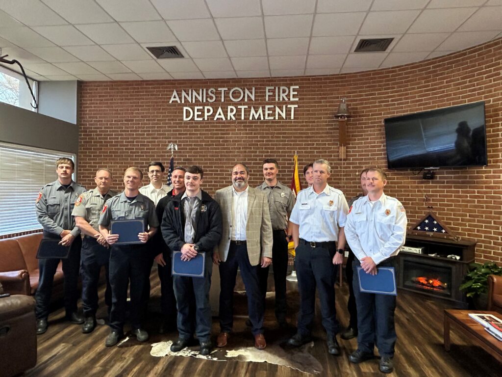 Photo of Anniston firefighters in uniform standing under a sign that reads "Anniston Fire Department" on a red brick wall.