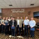 Photo of Anniston firefighters in uniform standing under a sign that reads "Anniston Fire Department" on a red brick wall.